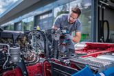 A person wearing safety equipment uses a cordless impact wrench on a vehicle chassis.