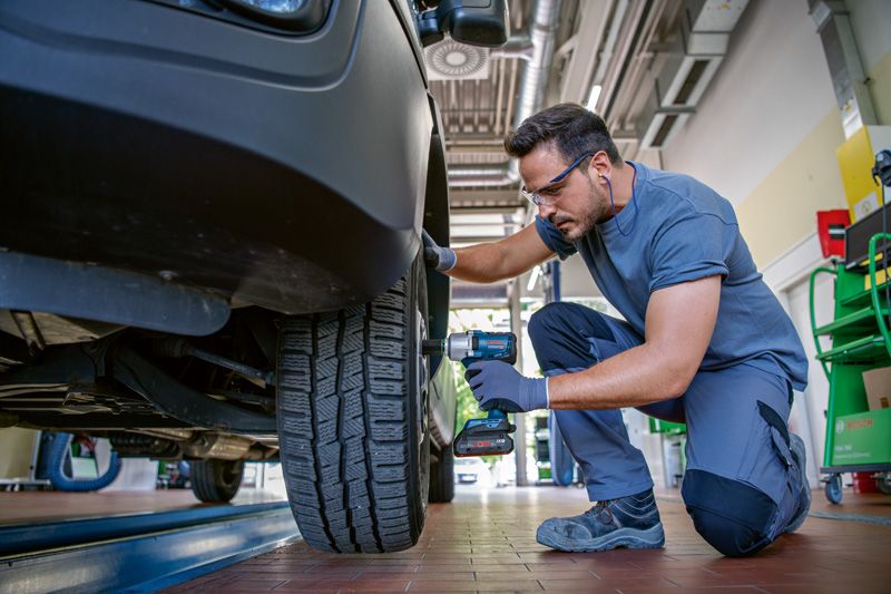 A person wearing safety equipment uses a cordless impact wrench to tighten a car wheel bolt.