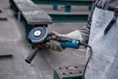 A person wearing safety equipment holds an angle grinder in an industrial setting.