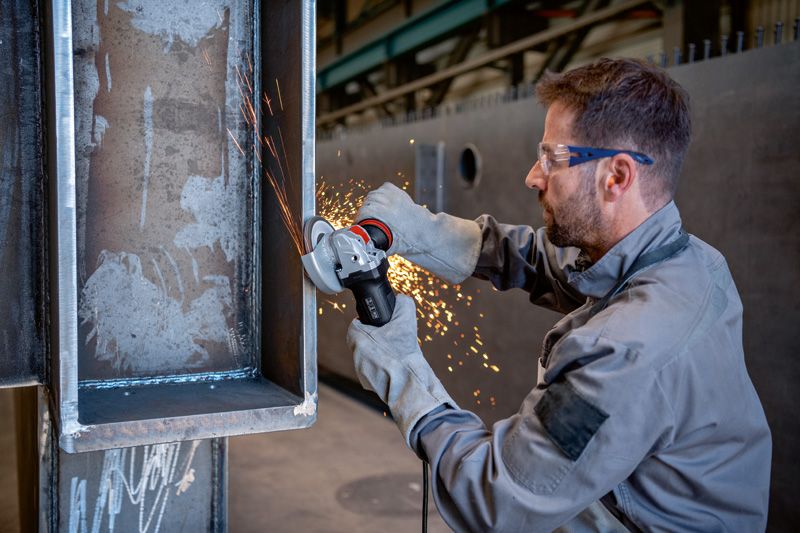 A person wearing safety equipment grinds a metal beam with an angle grinder, sparks flying.
