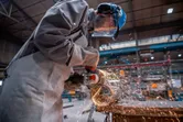A person wearing safety equipment grinds metal with an angle grinder, producing sparks.