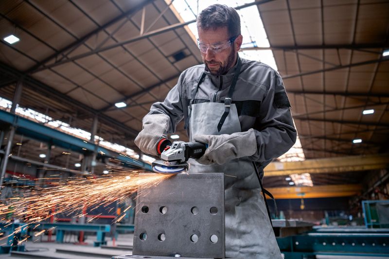 A person wearing safety equipment grinds a metal block, producing sparks in a factory.