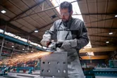 A person wearing safety equipment grinds a metal block, producing sparks in a factory.