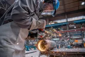 A person wearing safety equipment grinds a metal pipe with an angle grinder.