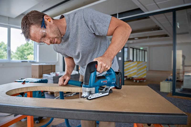 A person wearing safety equipment cuts a curved shape in wood with a cordless jigsaw.