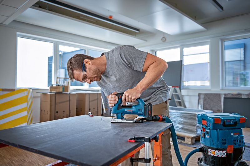 A person wearing safety equipment uses a cordless jigsaw to cut a large board indoors.