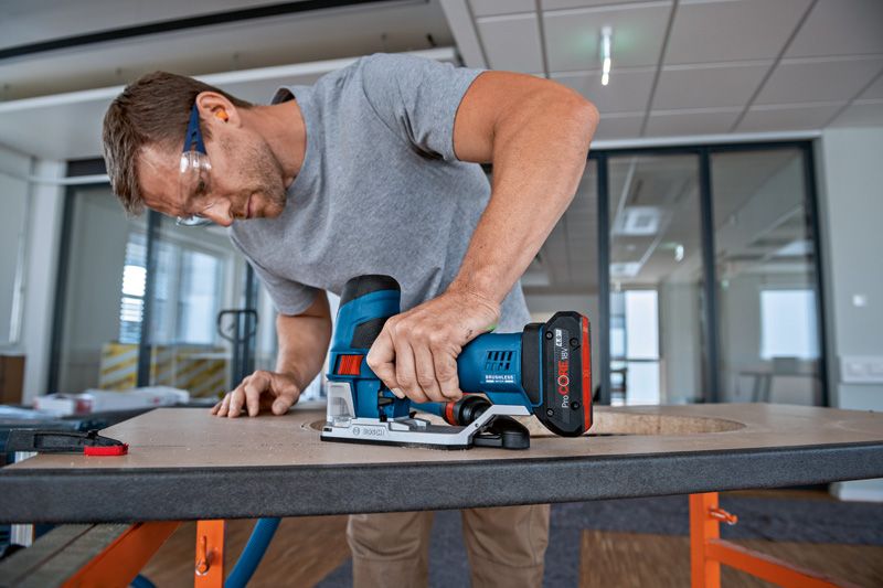 A person wearing safety equipment uses a cordless jigsaw to cut a hole in a wooden board.