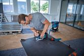 A person wearing safety equipment uses a jigsaw to cut a sheet on a workbench.