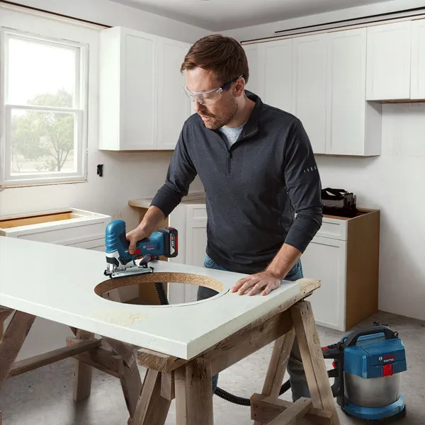 A person wearing safety equipment cuts a circular hole in a countertop with a cordless jigsaw.