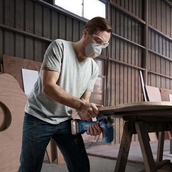 A person wearing safety equipment uses a cordless jigsaw to cut wood on a workbench.