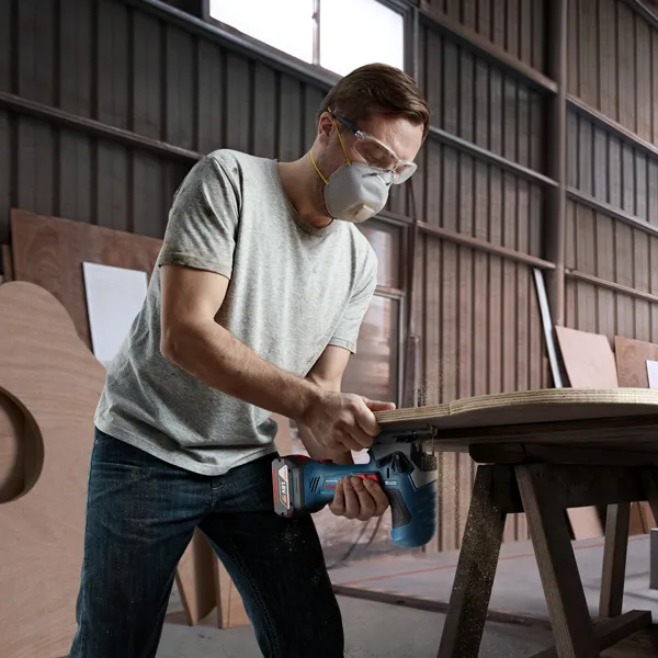 A person wearing safety equipment uses a cordless jigsaw to cut wood on a workbench.