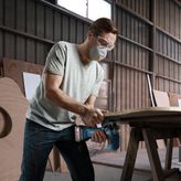 A person wearing safety equipment uses a cordless jigsaw to cut wood on a workbench.