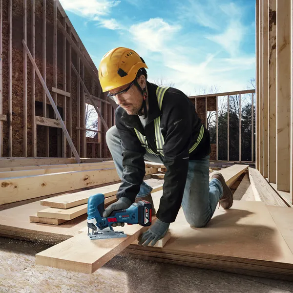 A person wearing safety equipment uses a cordless jigsaw to cut a wooden board.