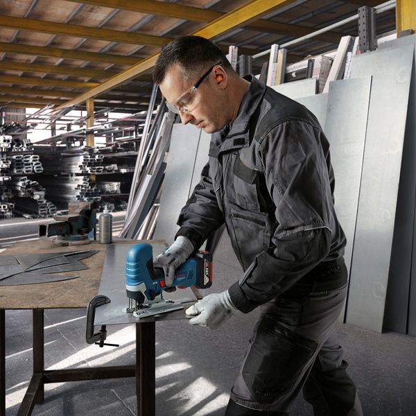A person wearing safety equipment uses a cordless jigsaw to cut a metal sheet.