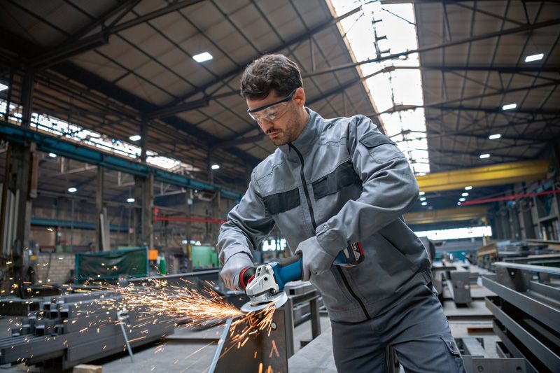GWS 18V-10 P A person wearing safety equipment grinds metal, creating sparks in an industrial workshop.