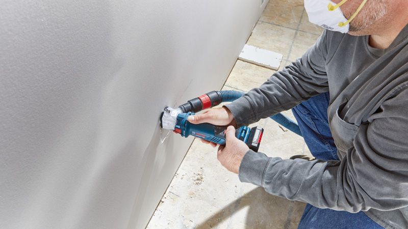 GCU 18V-30 A person wearing safety equipment cuts a hole in drywall using a cordless drywall router.
