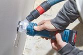 A person cuts a rectangular hole in drywall using a power tool with dust extraction.