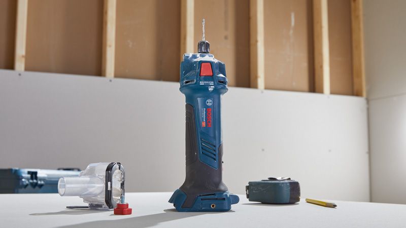 Cordless drywall router standing upright on a table next to its accessories and a pencil.