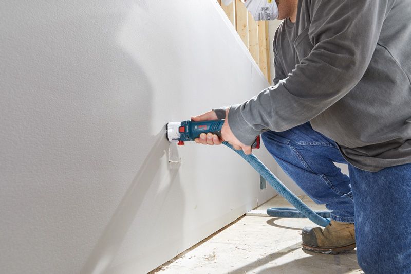A person wearing safety equipment uses a cordless drywall router to cut an opening.