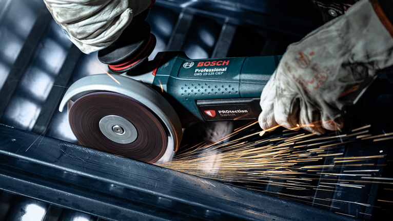 EXPERT N477 SCM Disc for Angle Grinders 125 x 22 mm, Extra Coarse Person wearing safety equipment uses an angle grinder to cut metal, producing sparks.