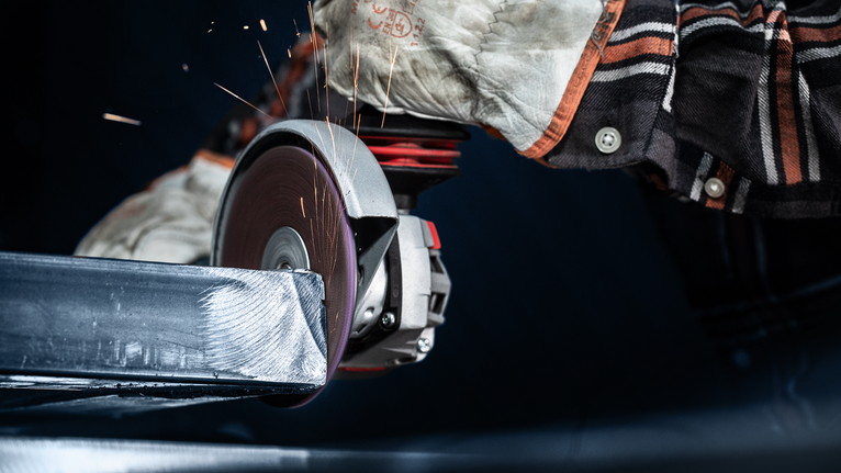 EXPERT N477 SCM Disc for Angle Grinders 125 x 22 mm, Extra Coarse Person wearing safety equipment grinds a metal beam with a power tool.