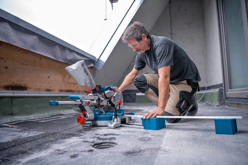 A person wearing safety equipment uses a cordless mitre saw to cut a long board.