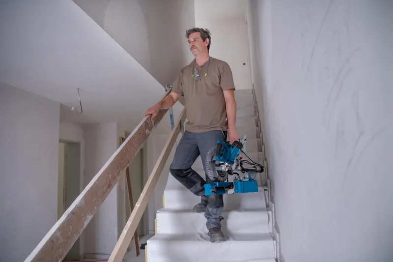 A person carries a mitre saw up a staircase inside an unfinished building.