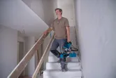 A person carries a mitre saw up a staircase inside an unfinished building.