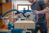 A person wearing safety equipment operates a mitre saw to cut a wooden beam.