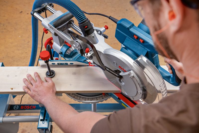 A person wearing safety equipment cuts wood with a mitre saw on a workbench.