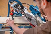 A person wearing safety equipment cuts wood with a mitre saw on a workbench.