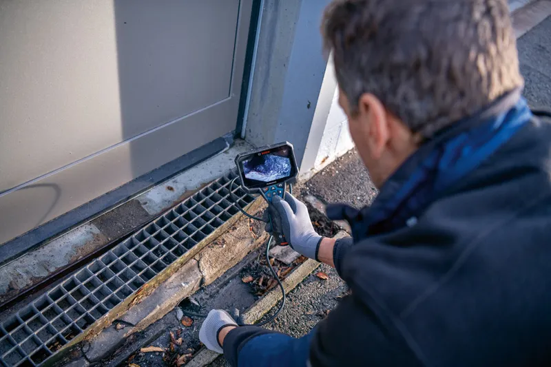 A person wearing safety equipment inspects a drainage grate using an inspection camera.