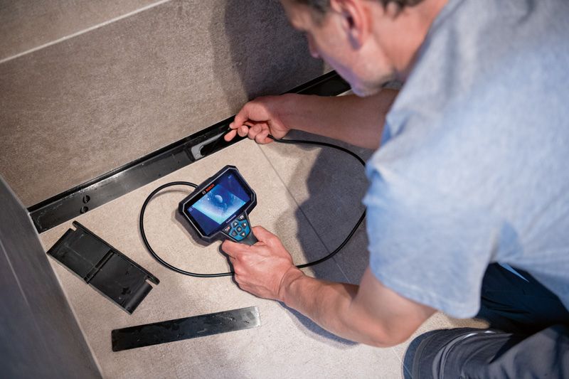 A person uses an inspection camera to check behind a panel on a tiled wall.