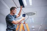 A person on a ladder inspects a ceiling panel with an inspection camera.