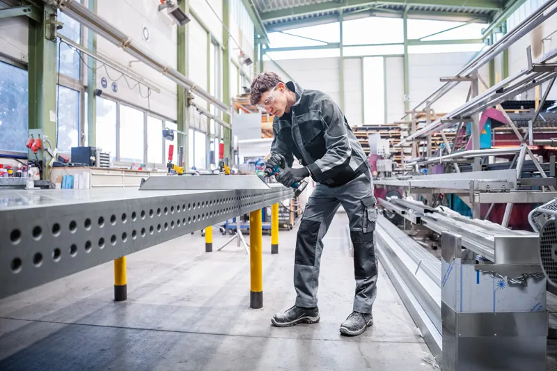 A person wearing safety equipment grinds metal edges on a perforated beam in a workshop.