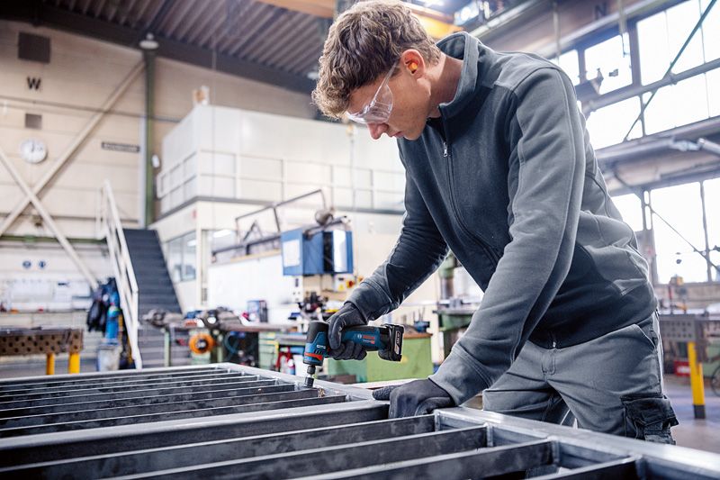 A person wearing safety equipment uses a rotary grinder on a metal frame.