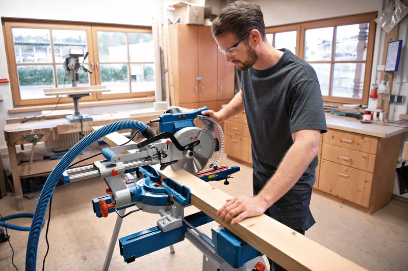 A person wearing safety equipment operates a mitre saw to cut a wooden plank.