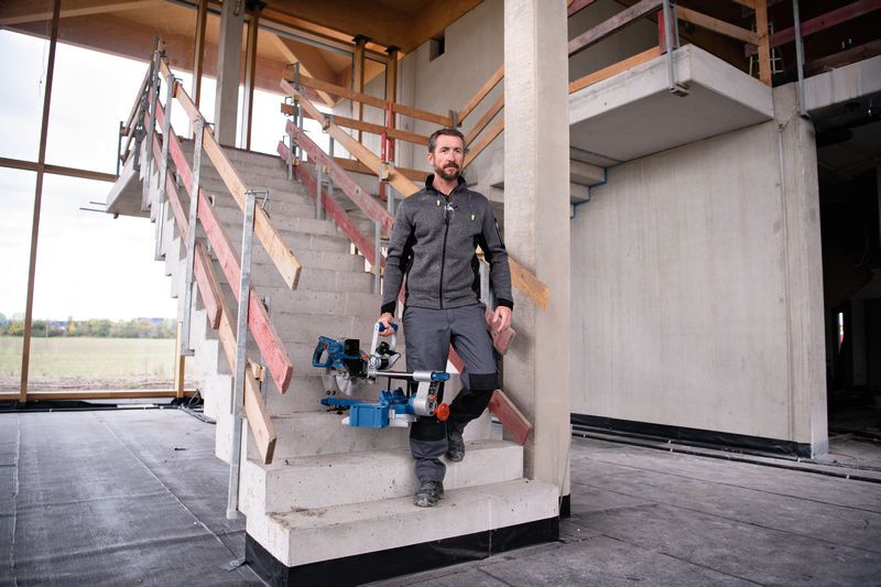 A person wearing safety equipment carries a cordless mitre saw down a staircase.