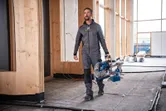 A person wearing safety equipment carries a cordless mitre saw at a construction site.