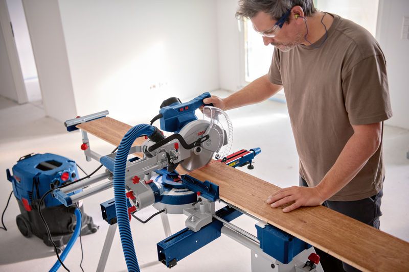 A person wearing safety equipment cuts a wooden board with a mitre saw.