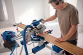 A person wearing safety equipment cuts a wooden board with a mitre saw.