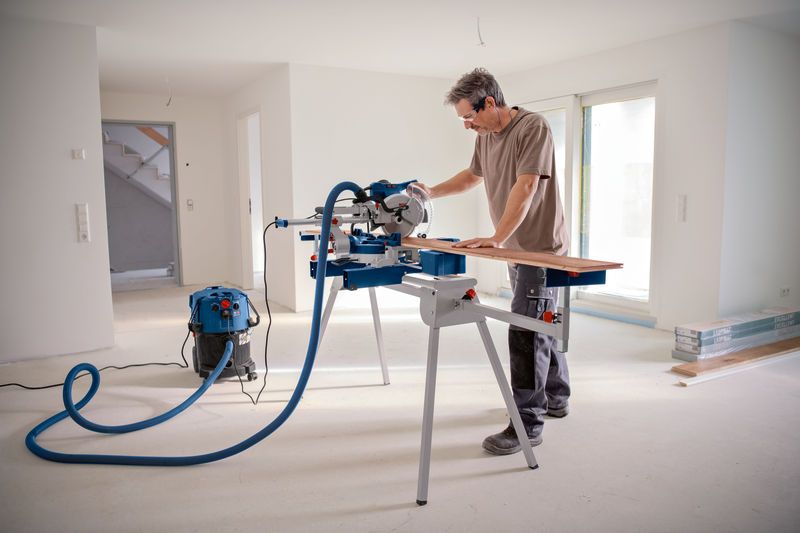 A person wearing safety equipment cuts wood on a mitre saw in a bright, unfinished room.