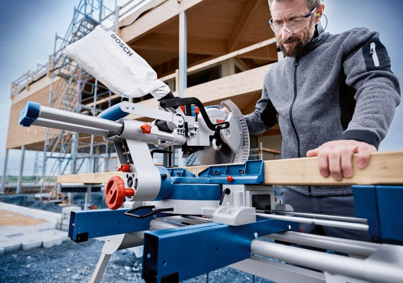 A person wearing safety equipment operates a cordless mitre saw to cut wood.