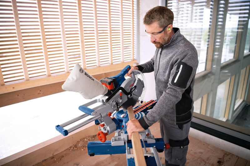 A person wearing safety equipment operates a cordless mitre saw to cut a wooden board.