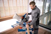 A person wearing safety equipment operates a cordless mitre saw to cut a wooden board.