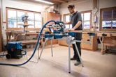 A person wearing safety equipment operates a mitre saw on a wooden board.