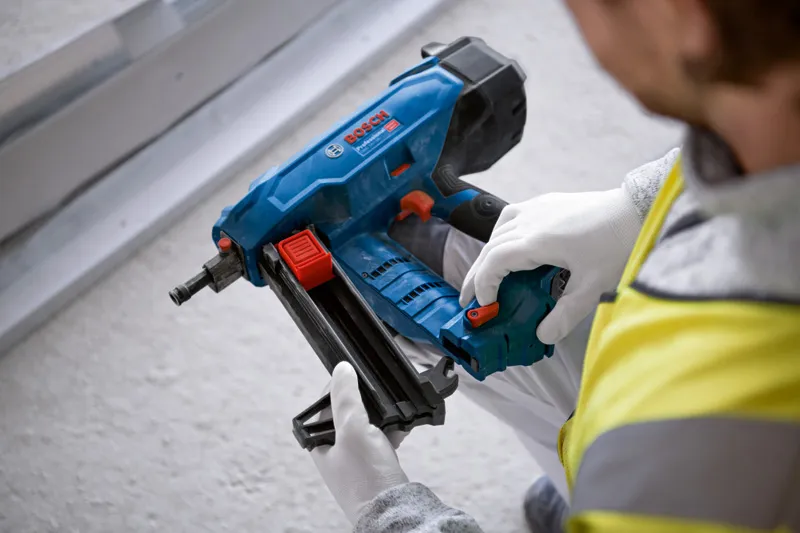 Person wearing safety equipment prepares a blue cordless nailer near a metal track.