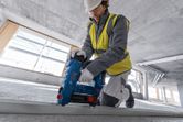 A person wearing safety equipment uses a cordless concrete nailer on a metal track.