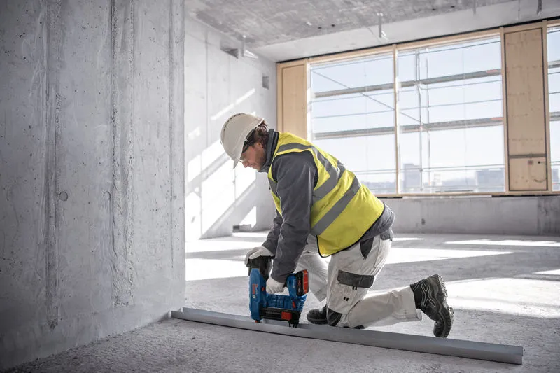 A person wearing safety equipment uses a power tool to fasten a metal track on a concrete floor.