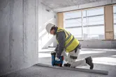 A person wearing safety equipment uses a power tool to fasten a metal track on a concrete floor.